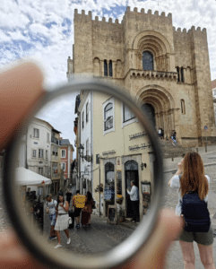 A person holds a circular lens that sharpens the view of a street scene in front of a historic stone church, with people walking and taking photos nearby.